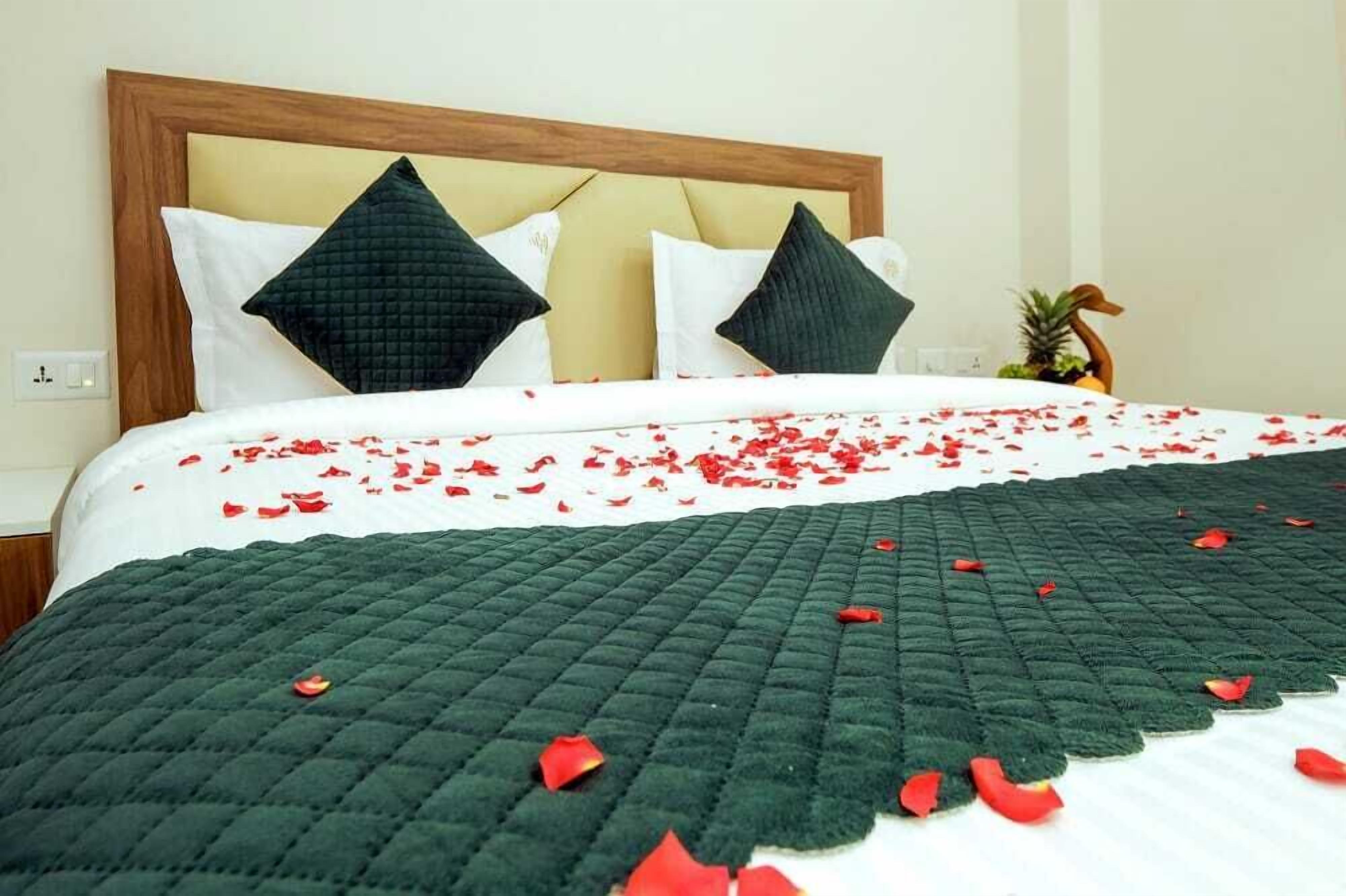 A neatly made bed with a green quilt, white linens, and red rose petals scattered across the bedspread.