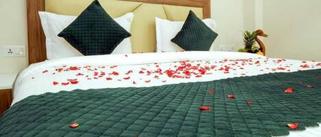 Una cama bien hecha con colcha verde, sábanas blancas y pétalos de rosa roja esparcidos sobre la colcha.
