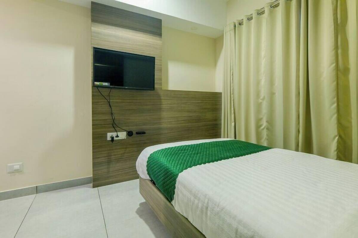 A hotel room with a bed, a flat-screen TV mounted on a wooden panel, and a curtain on the window.