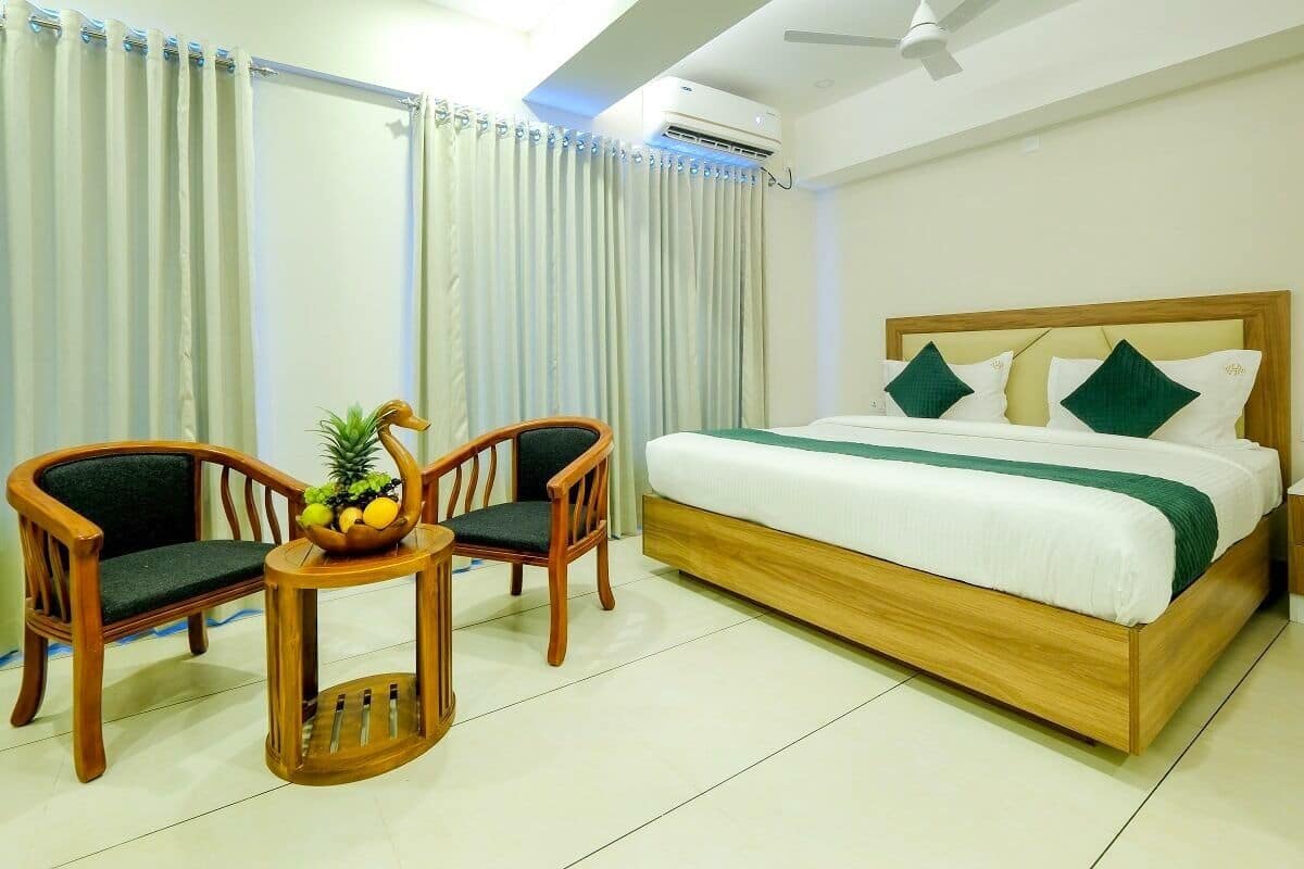 A bedroom with a wooden bed, two wooden chairs, a small round table with a fruit bowl, and a window with curtains.