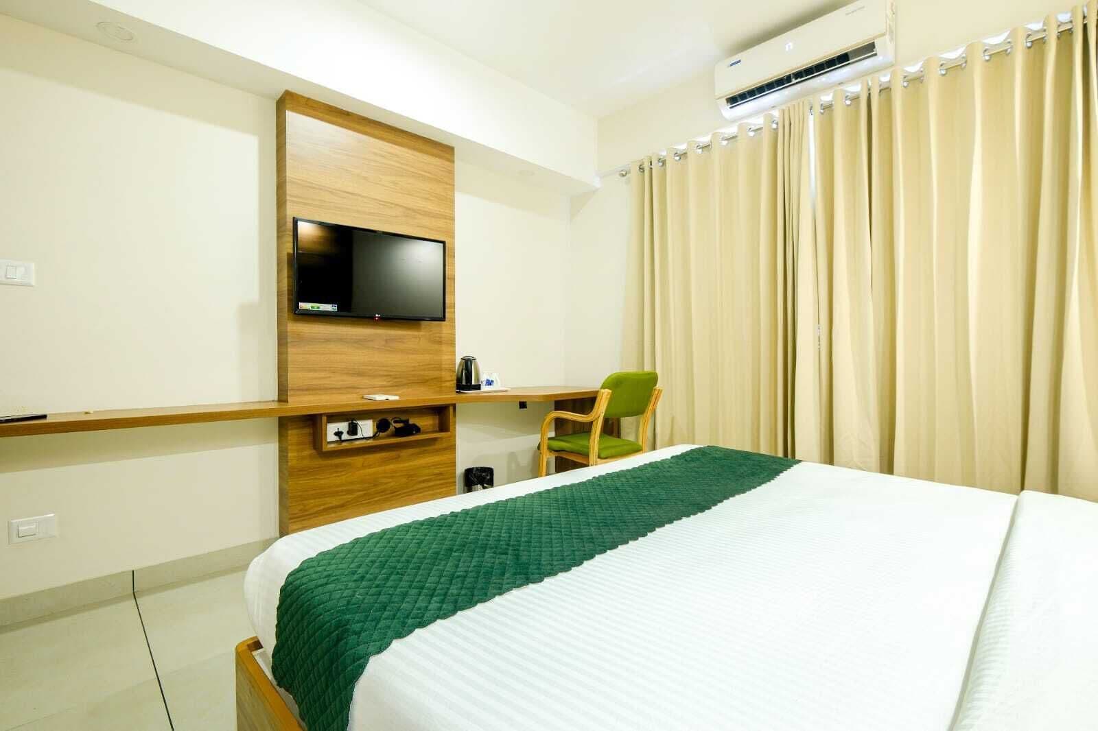 A hotel room with a bed, a TV mounted on a wooden panel, a chair, and a curtain.