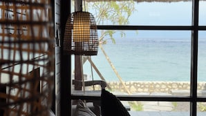 A room with a large window overlooking the sea, a wicker lamp, and a thatched roof visible through the window.