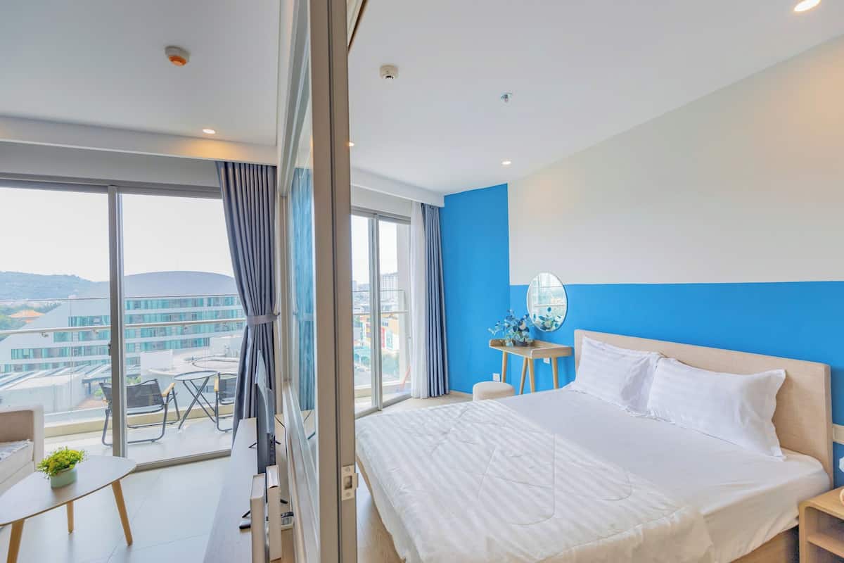 A modern hotel room with a large bed, a desk, and a view of the city through large windows.