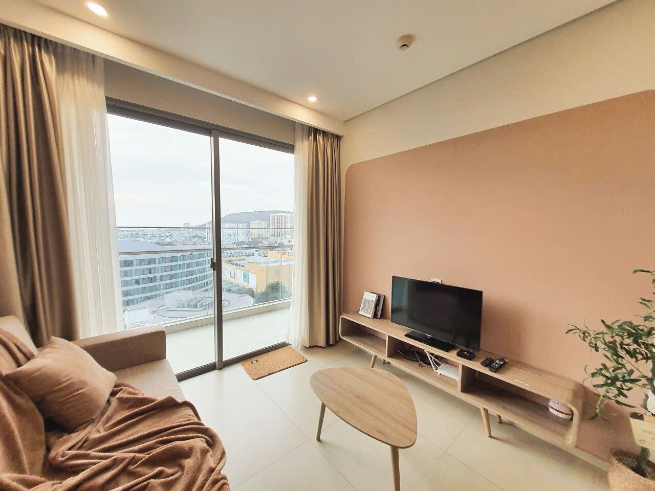Un salon moderne avec une grande fenêtre, un canapé, un téléviseur posé sur un meuble en bois et une petite table.