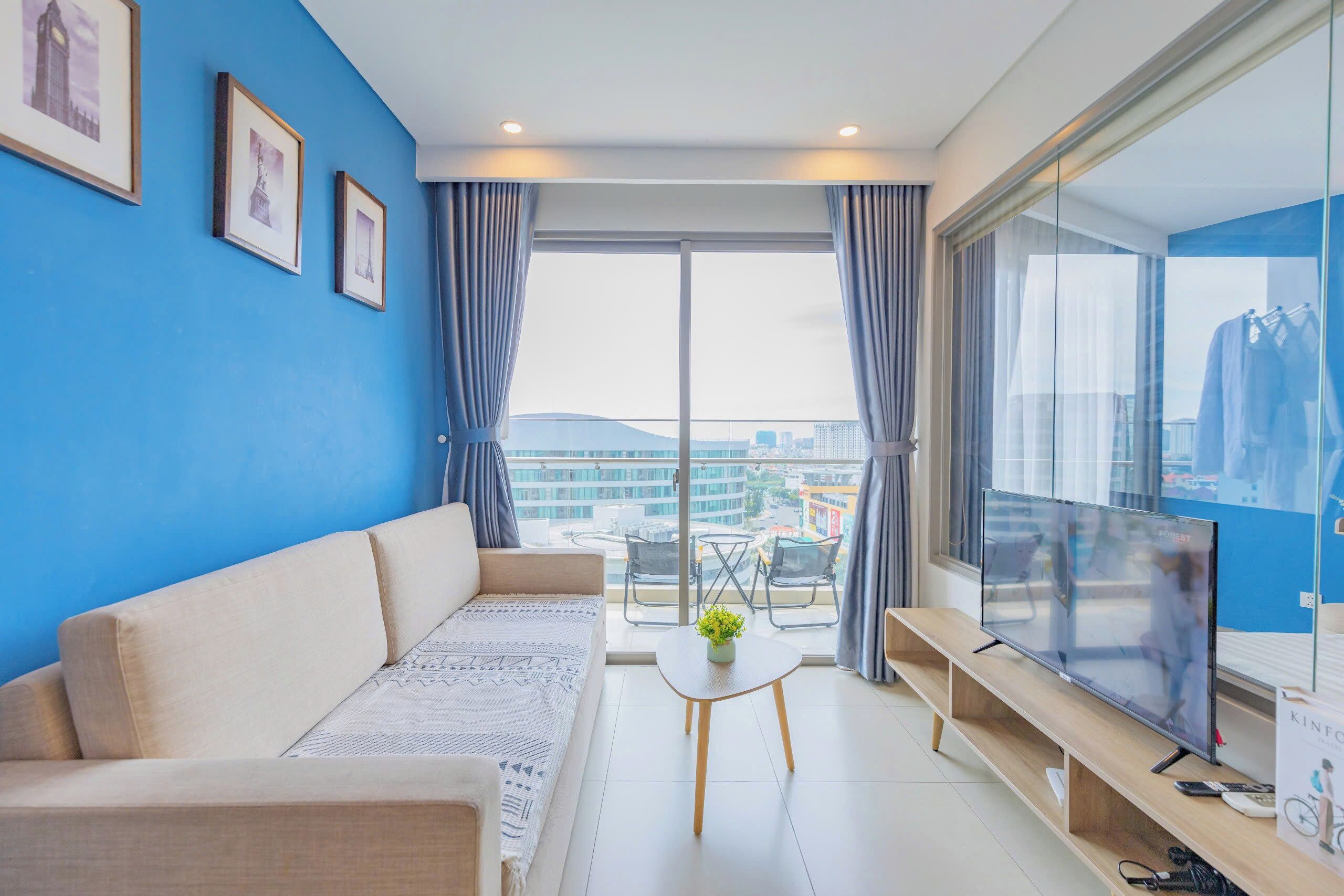 A modern living room with a sofa, a coffee table with a plant, a flat-screen TV on a stand, and a balcony with a view of the city.