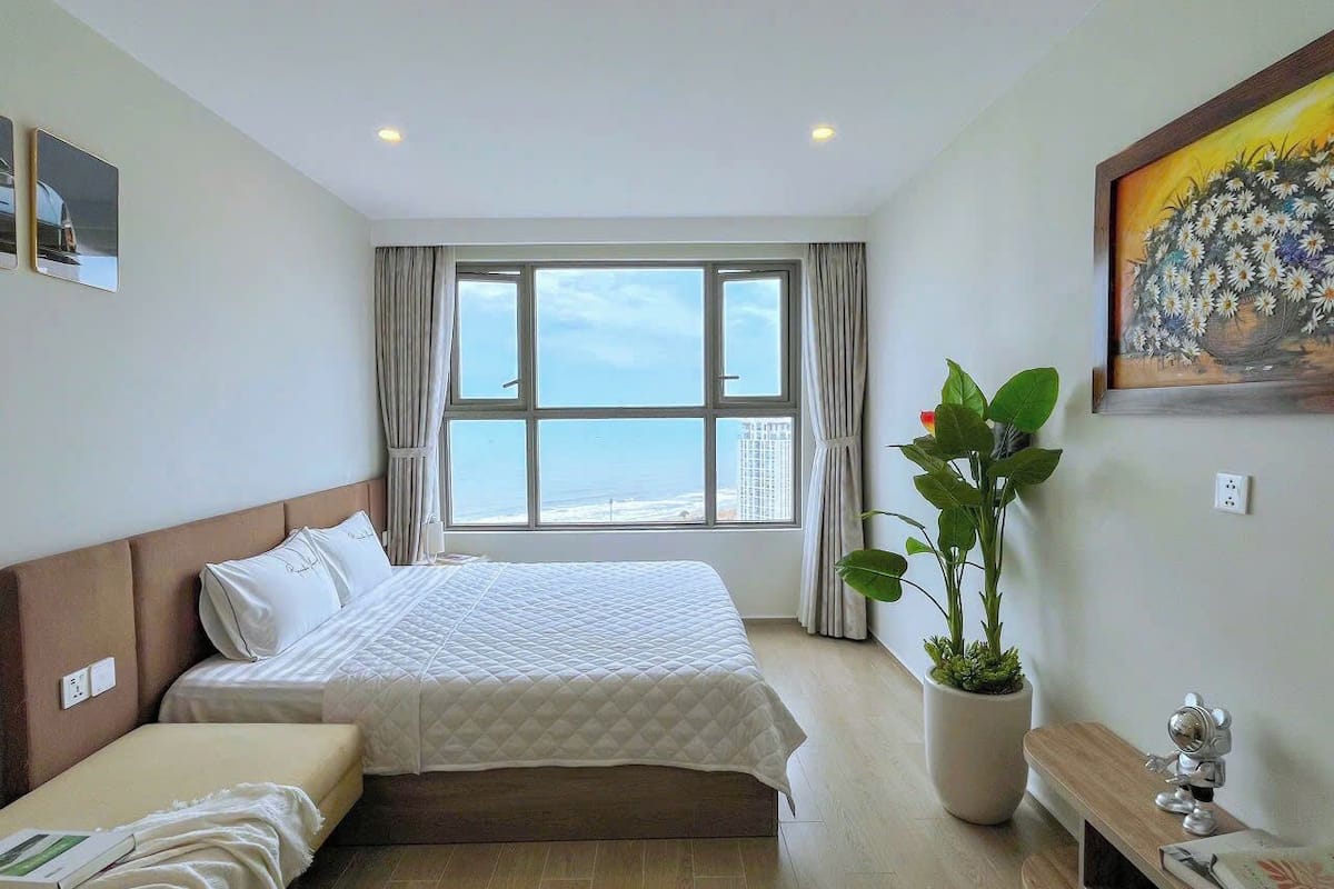 A hotel room with a large bed, a bedside table, a potted plant, and a view of the ocean.
