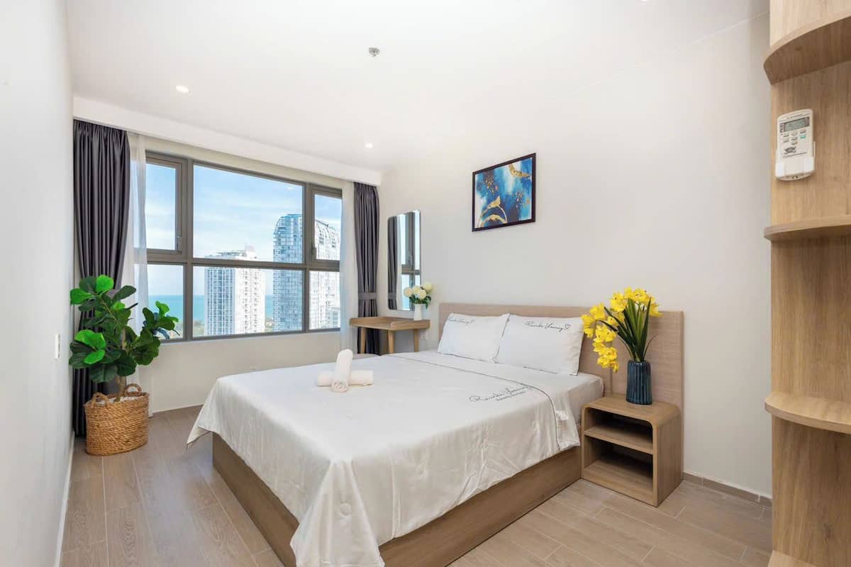 A modern hotel room with a large bed, a view of the city, a small table with a vase of flowers, and a wall-mounted thermostat.