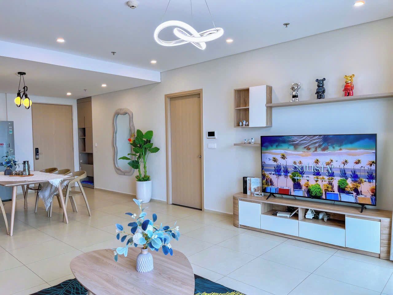 A modern living room with a flat-screen TV, a dining area with a table and chairs, and a large potted plant.