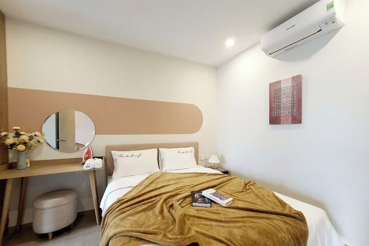 A hotel room with a bed, a desk, a chair, a mirror, a wall-mounted air conditioner, and a small trash bin.
