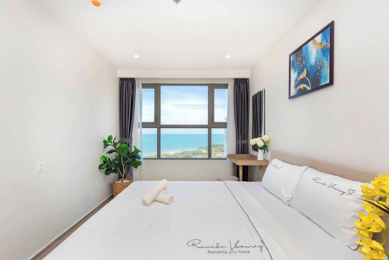 A hotel room with a bed, a desk, a chair, a plant, and a painting.