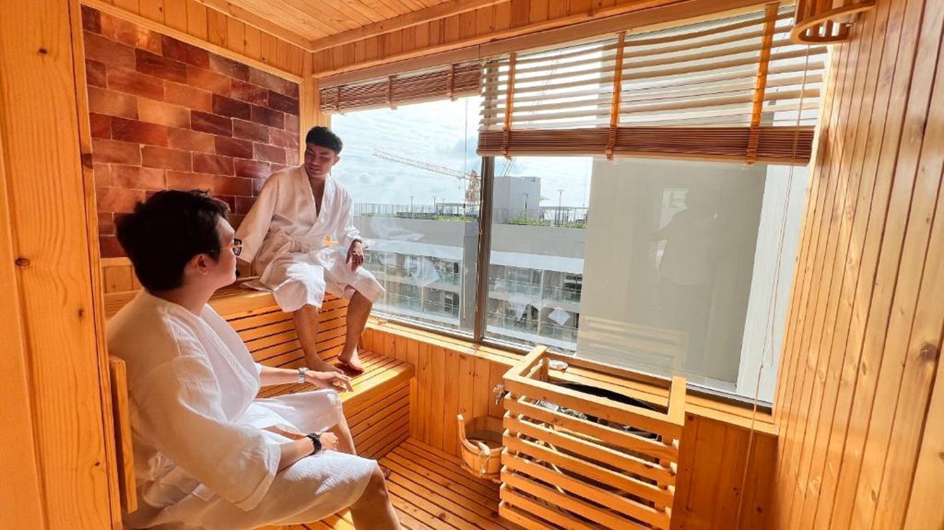 A wooden sauna room with a brick wall, wooden benches, and a large window with blinds.