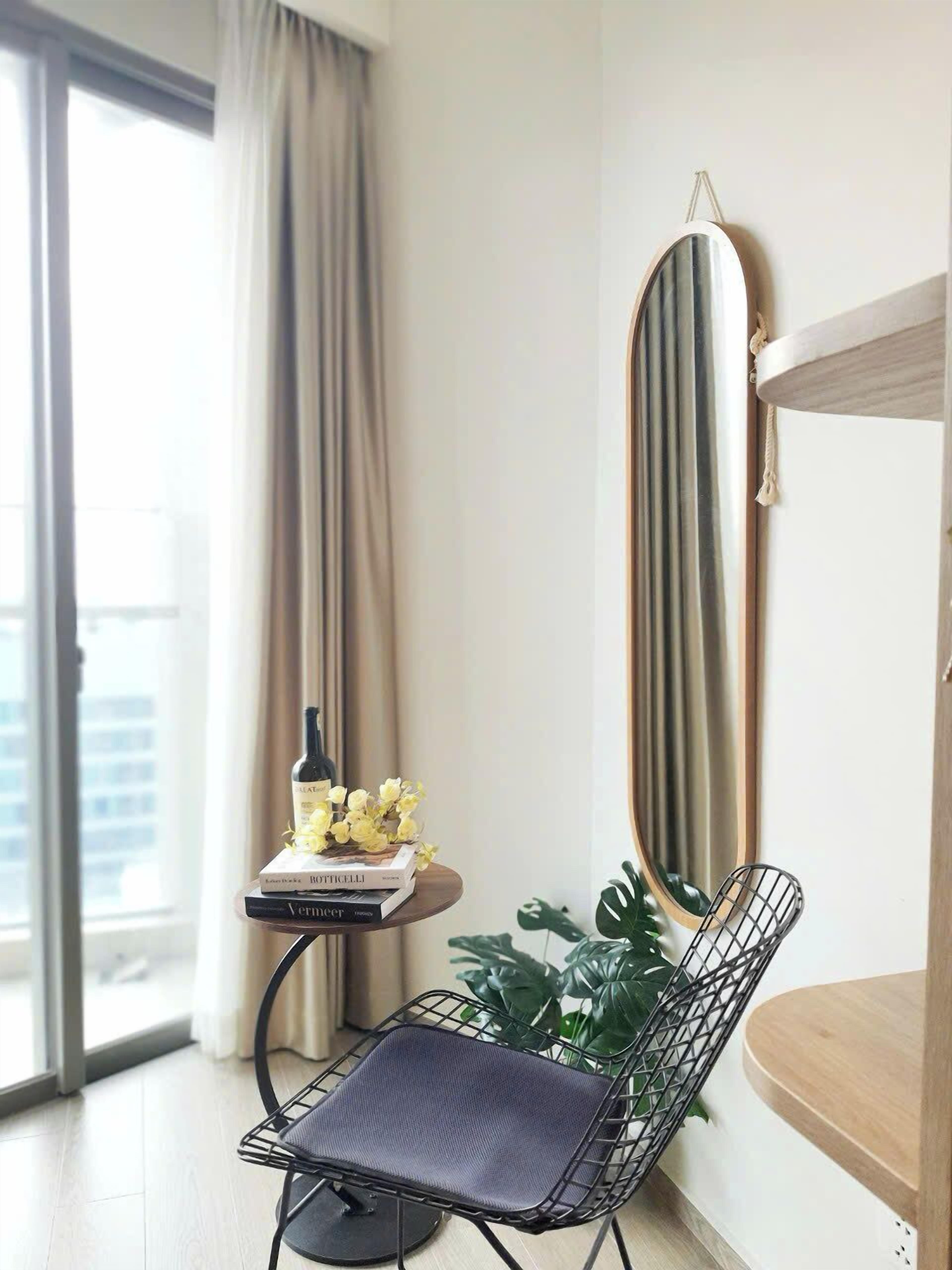 Una sala de estar moderna con una silla de alambre negra, una mesita con una botella y libros, un espejo grande y una planta en maceta.