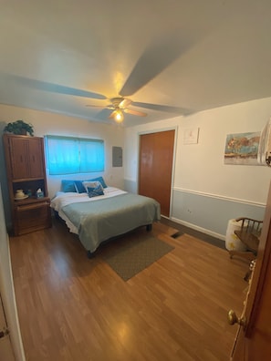 Ein Schlafzimmer mit einem Bett, einem Holzschrank, einem Deckenventilator, einem Fenster mit Jalousien und einer Tür.