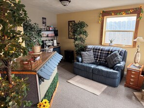 A cozy living room with a blue sofa, a wooden side table, a small rug, and a window with a view.