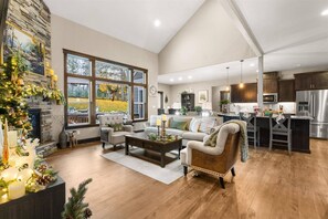 A spacious living room with a fireplace, large windows, and a kitchen area with dark wooden cabinets and stainless steel appliances. - Four Bedroom Retreat Near Hayden Lake: Game Room, Dog-Friendly, Ping Pong Table, Long Stays Welcome (Coeur d'Alene)