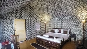 A bedroom with a floral-patterned ceiling, a wooden bed, a nightstand, a chair, and a desk with a lamp. - DESERT LEELA RESORT (Jaisalmer)