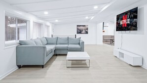 Ein modernes Wohnzimmer mit einem grauen Ecksofa, einem weißen Couchtisch und einem an der Wand befestigten Fernseher mit dem Netflix-Logo.