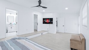 Ein geräumiges Wohnzimmer mit einem großen, an der Wand befestigten Fernseher, einem Deckenventilator und einem beigefarbenen Sessel.