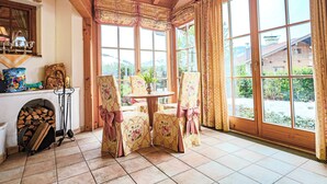 Ein gemütlicher Winkel mit Kamin, Tisch und Stühlen, großen Fenstern mit Vorhängen und Blick auf ein Gebäude.