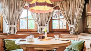 Ein gemütlicher Raum mit einem runden Holztisch, grünen Sitzkissen und großen Fenstern mit Vorhängen.