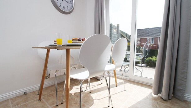 A dining area with a table and two white chairs, a clock on the wall, and a view of the outside through a large window. - Gwent View by Tŷ SA (Newport)