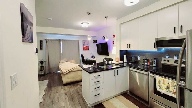 A modern kitchen with white cabinets, a stainless steel refrigerator, and a microwave. There is a dining area with a table and chairs, and a small kitchenette with a stove and sink. - Cozy Condo-Apartment/Suite in Brampton (Brampton)
