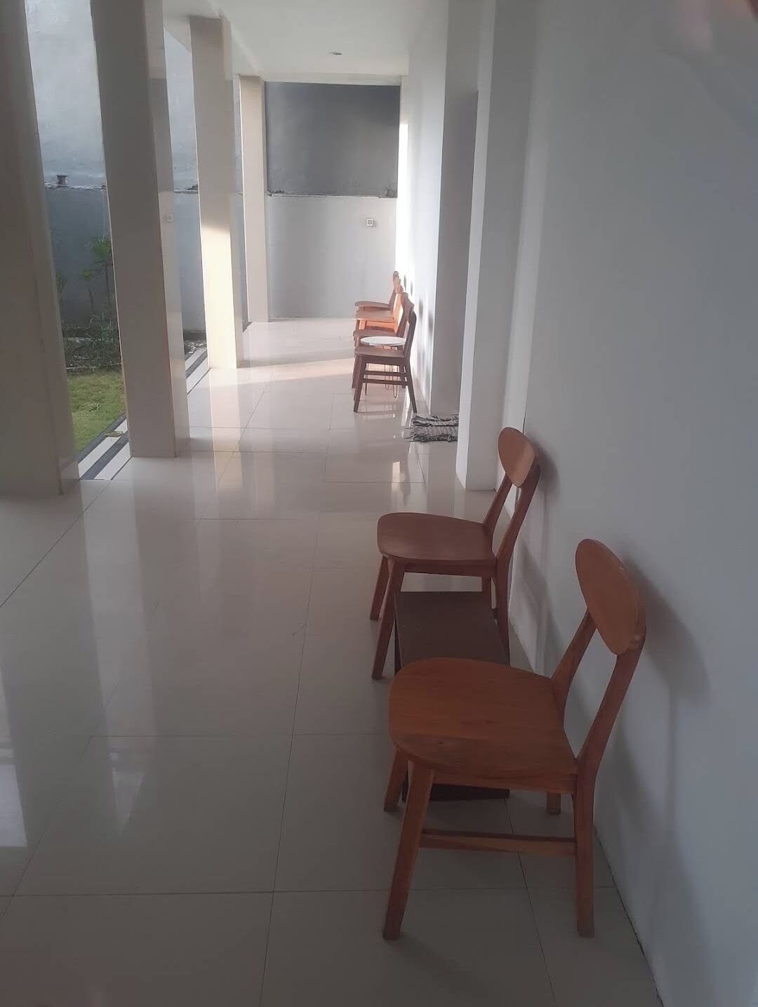 Un couloir bordé de chaises en bois le long du mur, qui mène à une pièce dotée d’une grande fenêtre.