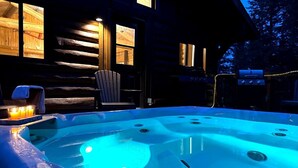 Un jacuzzi con luz azul, una silla de madera y una parrilla frente a una cabaña de troncos.