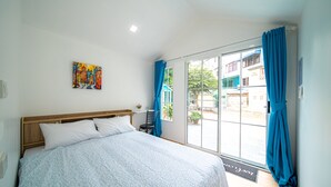 A bedroom with a bed, a chair, a window with curtains, and a door leading to an outdoor area. - UrbanNest Hostel  (Pattaya)