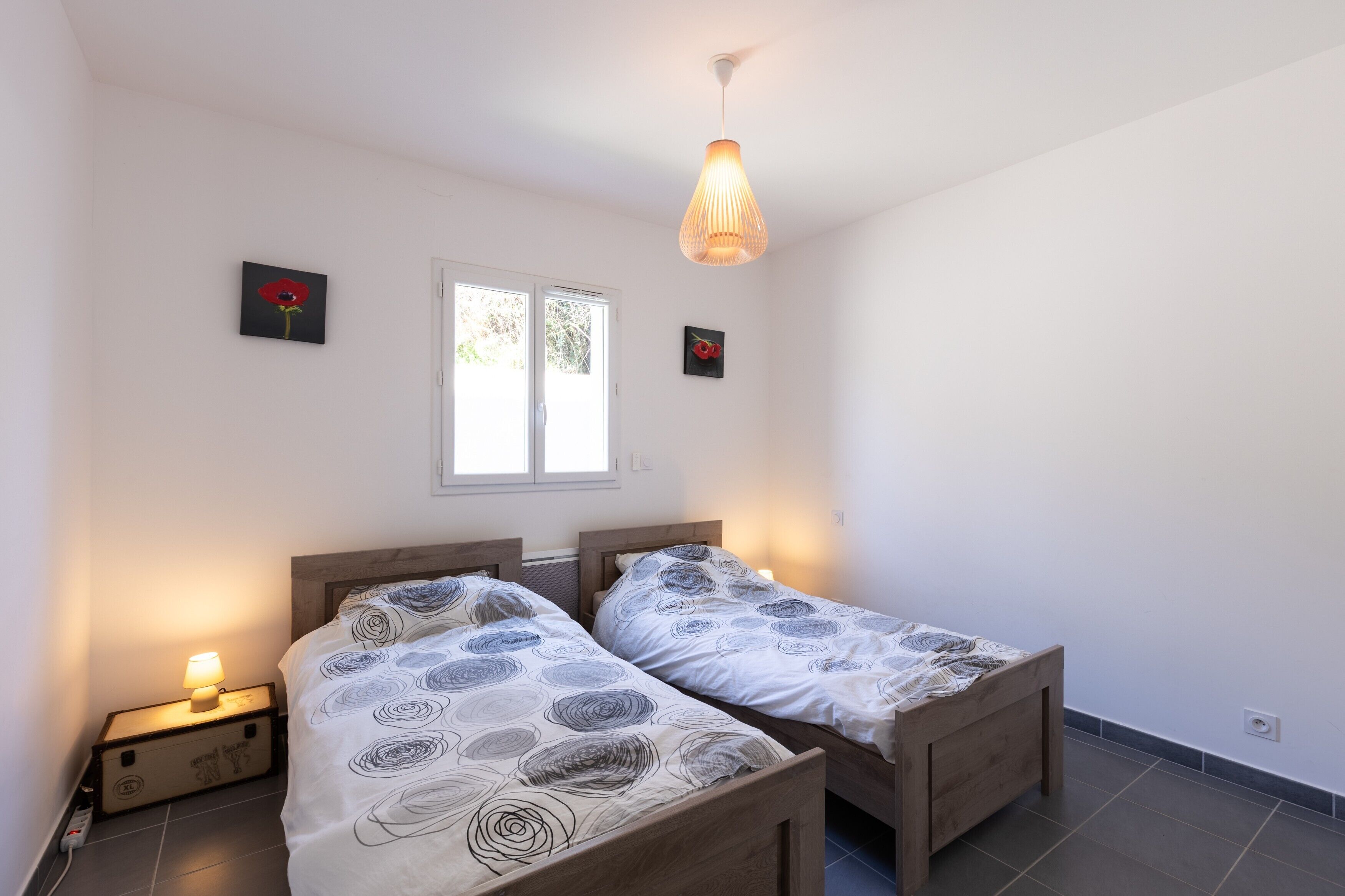 Une chambre avec deux lits simples, une table de chevet avec une lampe, une fenêtre et un luminaire suspendu.