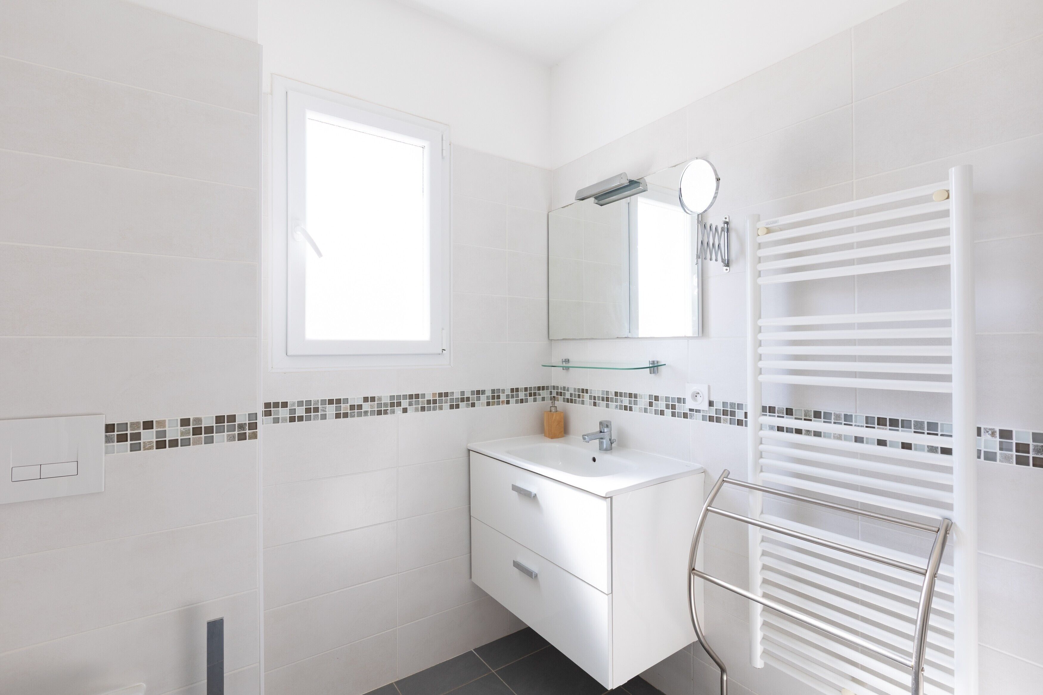 Bagno con piastrelle bianche, una finestra, uno specchio, un lavabo e un portascopevoli.