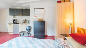 Una habitación pequeña con cama, escritorio, silla, armario y kitchenette.