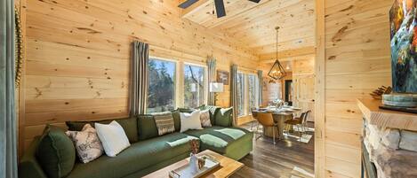 L’intérieur chaleureux d’un chalet avec un canapé vert, un plancher en bois, un coin repas et un ventilateur de plafond.