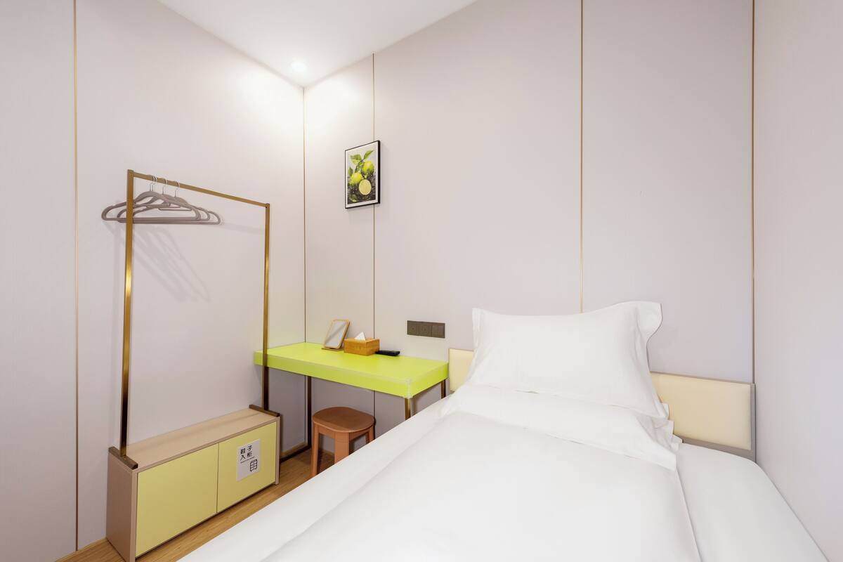 A hotel room with a bed, a desk with a chair, a small stool, a wardrobe, and a mirror.