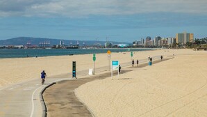 Ein Strand mit einem Radweg, einem Hinweisschild und einer in der Ferne liegenden Stadtlandschaft.