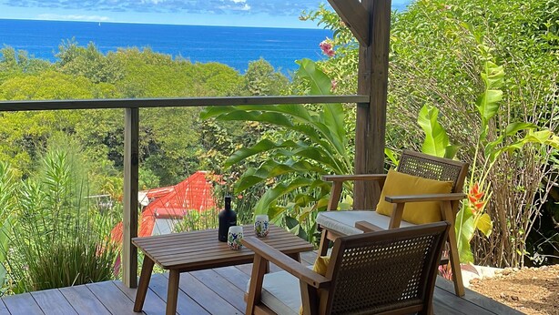 A wooden deck with a table and two chairs, overlooking a lush landscape and the ocean. - Charming T1 50 m from Anse Bonneville beach, beach and idleness guaranteed! (La Trinité)