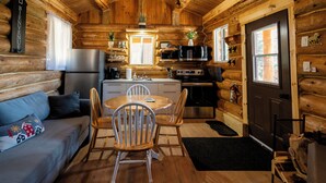 Interior de uma cabana de tronco com cozinha, sala de jantar e um sofá com almofada decorativa.