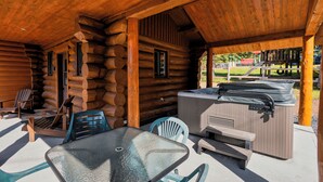 Uma cabana de toras com mesa e cadeiras de vidro, churrasqueira e banheira de hidromassagem.