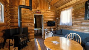 Interior de uma cabana de tronco com lareira, televisão, sofá e mesa de jantar.