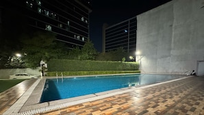 A swimming pool with a wooden deck, surrounded by a hedge and a building with multiple windows. - The Sandalwood By La Carnation (Sohna)