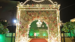An ornate archway with floral decorations and a chandelier, leading to a green hedge and a white fence. - The Sandalwood By La Carnation (Sohna)