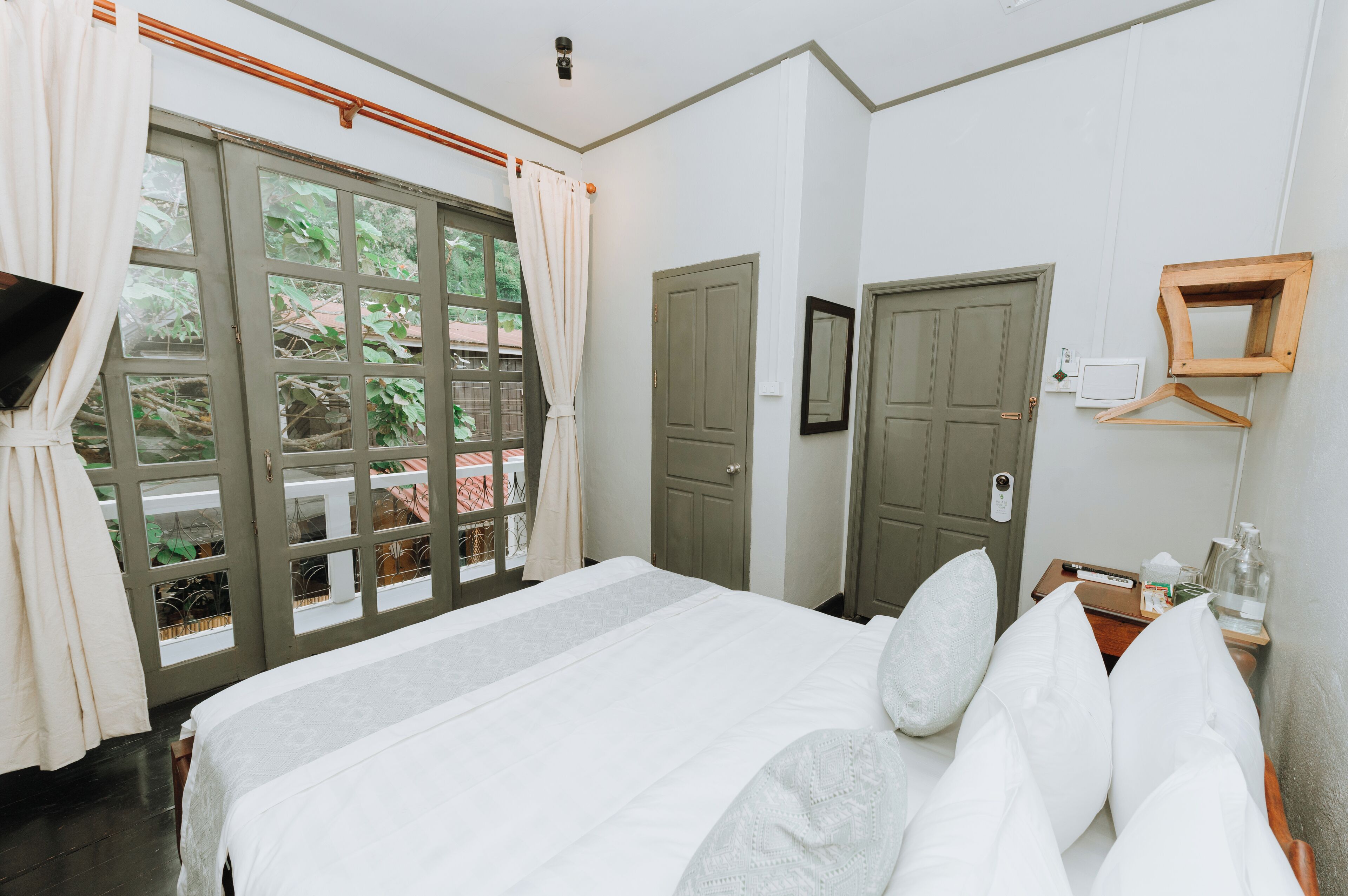 A bedroom with a large bed, a window with curtains, a door, a bedside table with a bottle and tissues, and a wall-mounted TV.