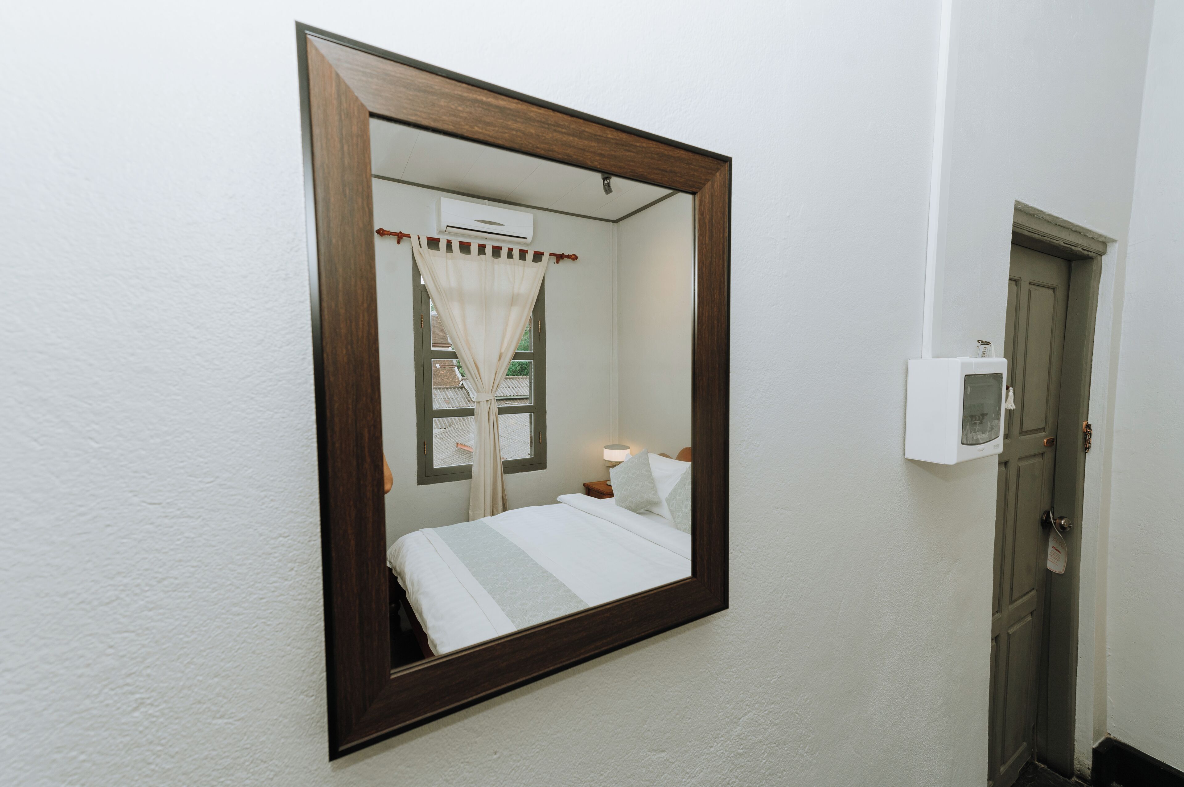 A bedroom with a large mirror, a bed with white bedding, a bedside table with a lamp, and a window with curtains.