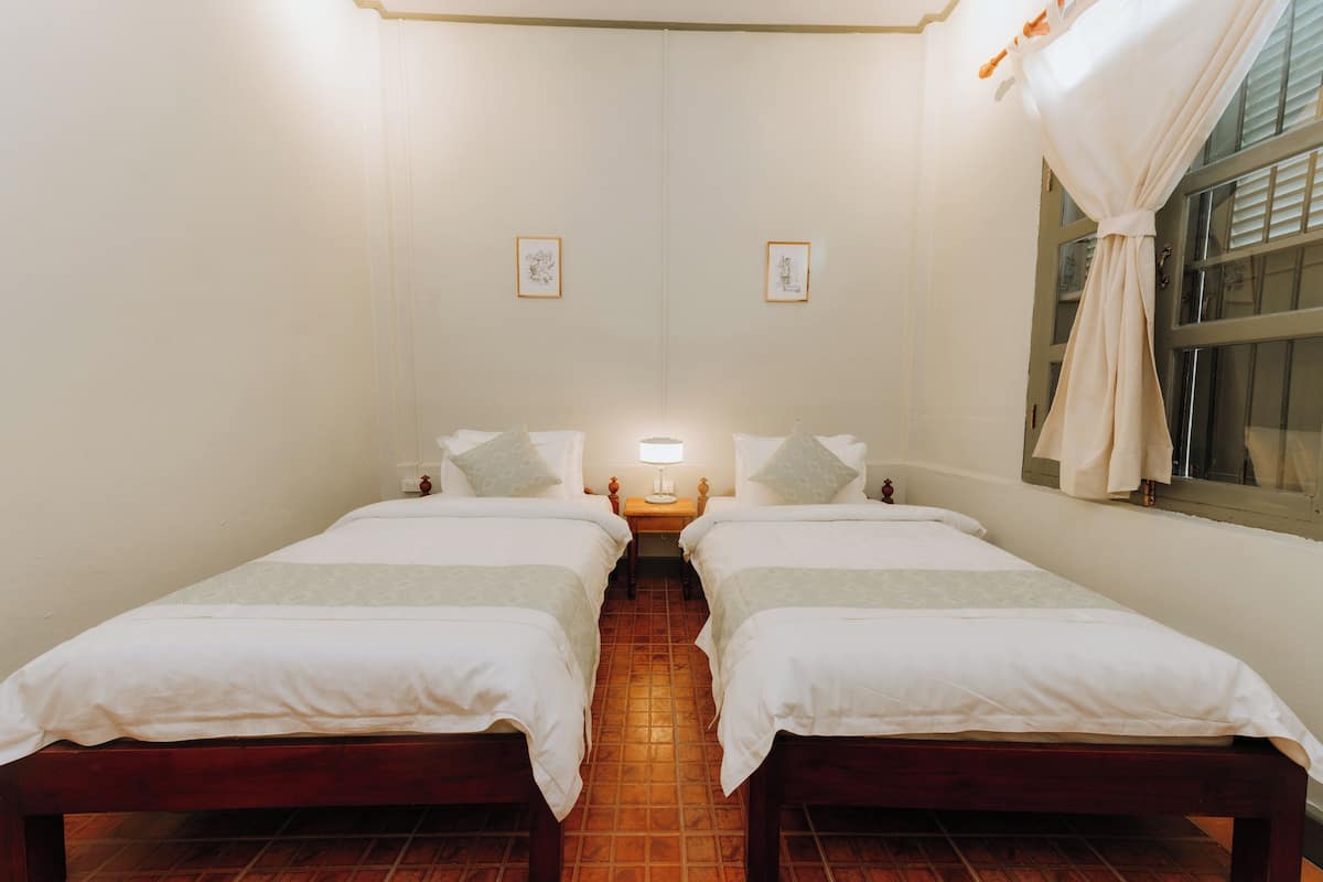 Quarto com duas camas de solteiro, uma mesa de cabeceira com luminária e uma janela com cortinas.