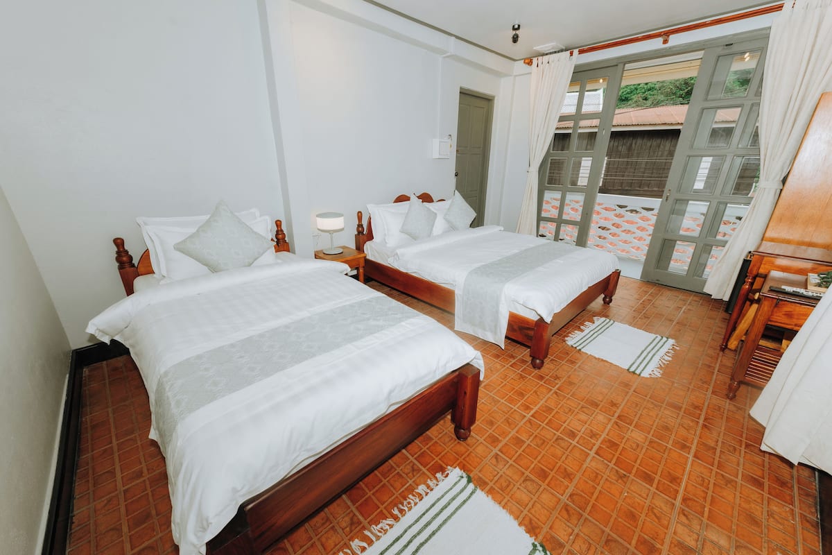 Quarto de hotel com duas camas de madeira, uma pequena mesa, um criado-mudo e vista para o exterior através de uma porta com painéis de vidro.