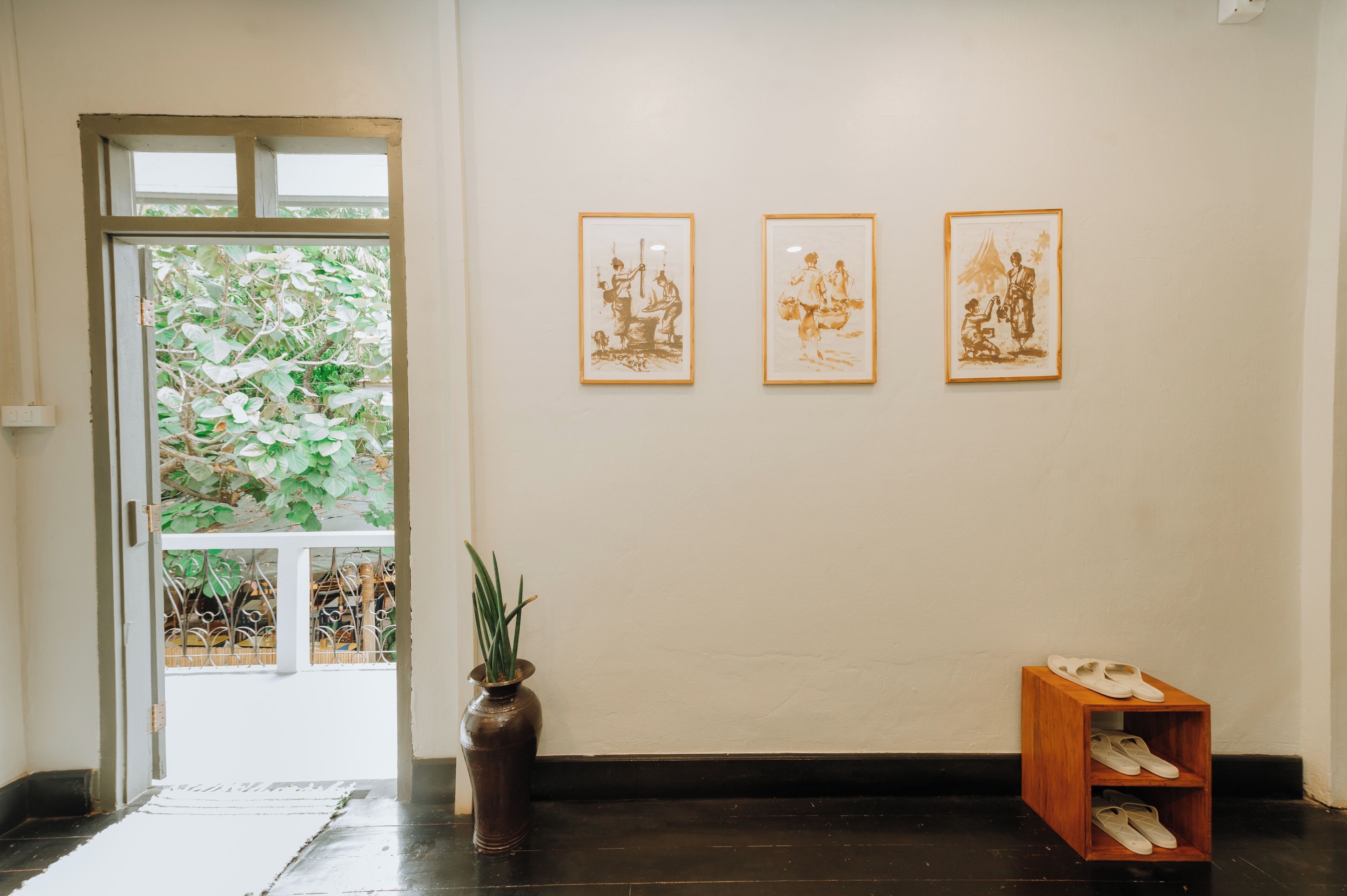 A hallway with a door leading to a balcony with plants, three framed artworks on the wall, a potted plant, and a shoe rack.