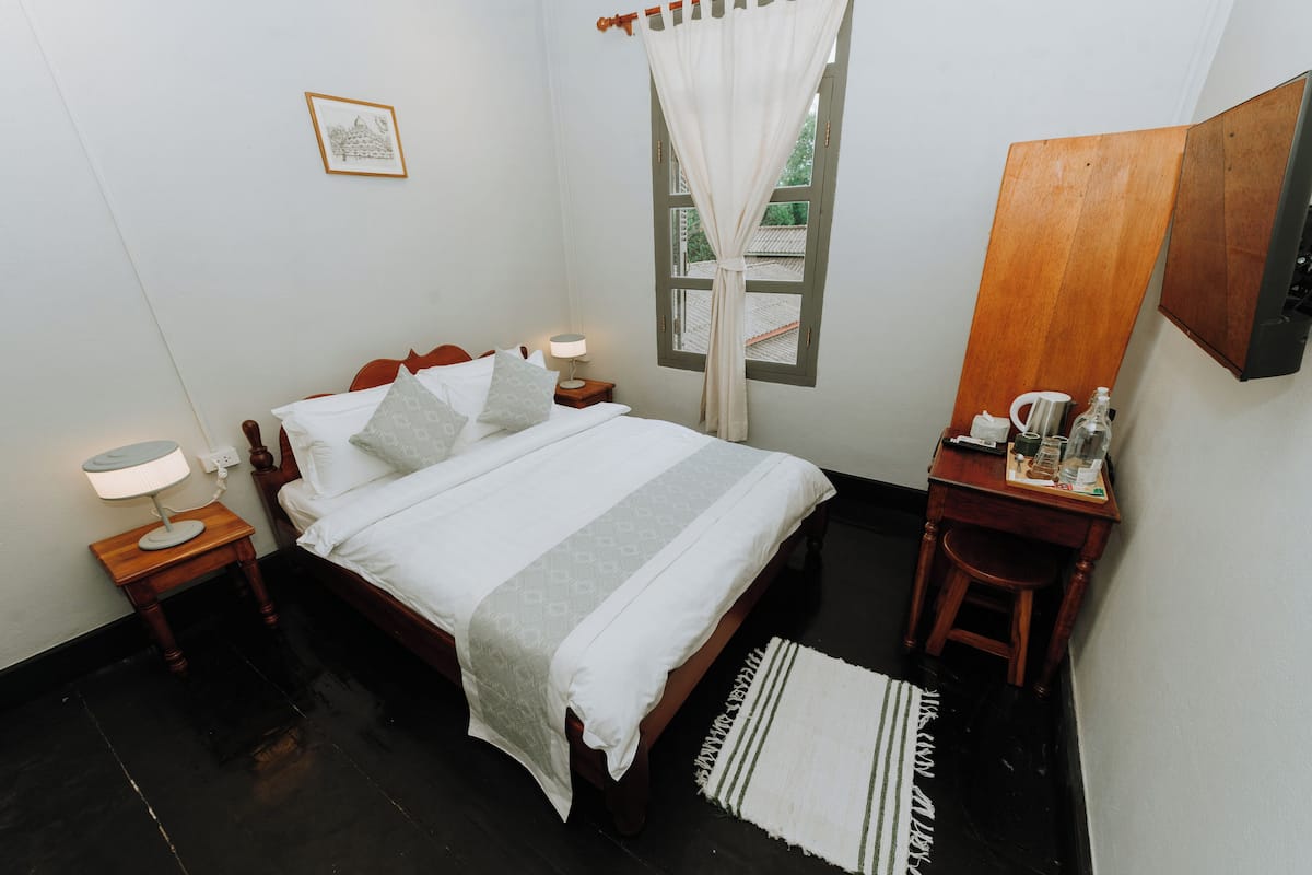 Quarto bem arrumado com cama, mesas de cabeceira, um criado-mudo, janela com cortinas, escrivaninha de madeira e uma cadeira.