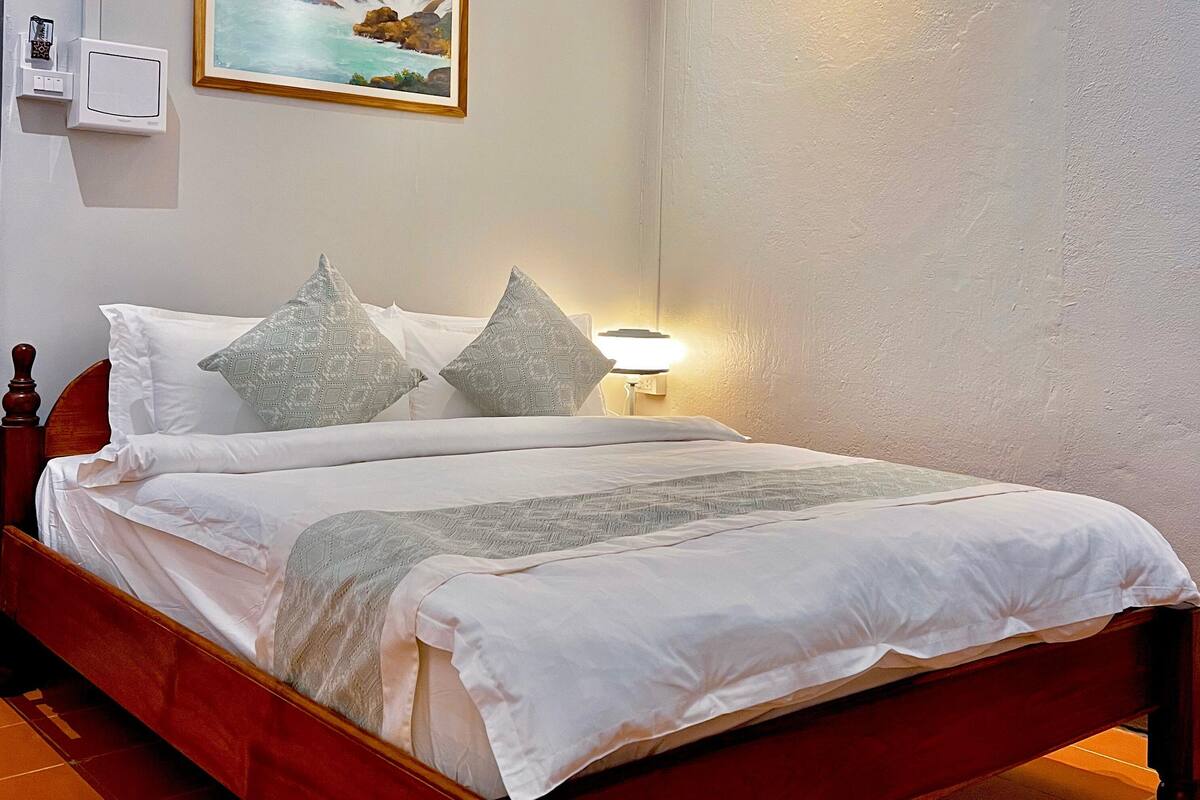 Uma cama bem arrumada com lençóis brancos e almofadas estampadas, um abajur e uma pintura de uma cascata na parede.