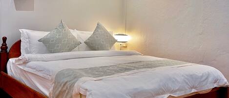 A neatly made bed with white linens and patterned pillows, a bedside lamp, and a painting of a waterfall on the wall.