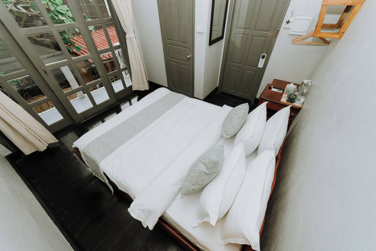 Cama bem arrumada com lençóis e almofadas brancas, mesa de cabeceira de madeira com uma garrafa e um copo, e porta com um espelho e uma pequena prateleira.